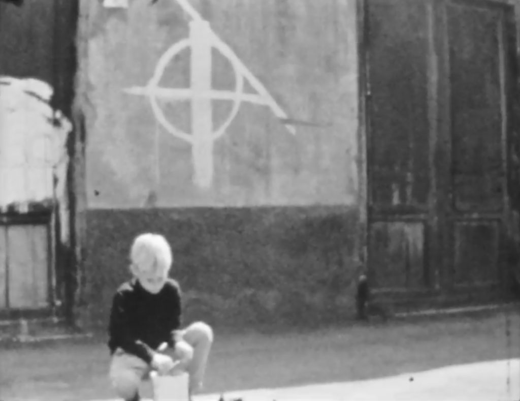 Boy on a street crouched, behind him a wall with a symbol on it